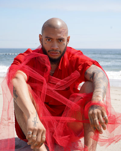 A model sitting in the sand at the beach dressed in red and wearing various Automic Gold jewelry