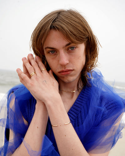 A model sitting in the sand at the beach dressed in blue and wearing various Automic Gold jewelry