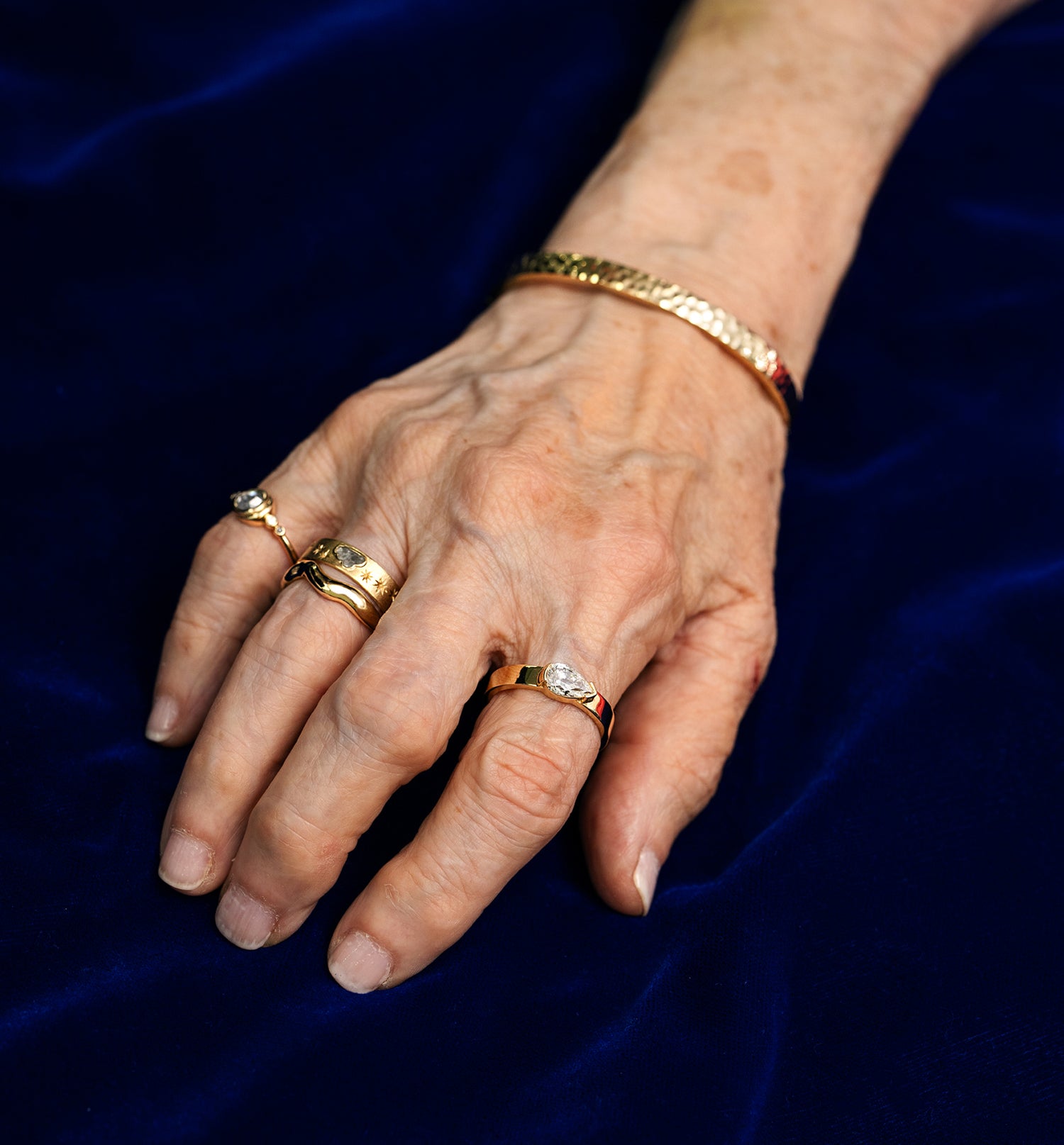 An older models hand wearing 4 different yellow gold rings and a yellow gold hammered bangle bracelet