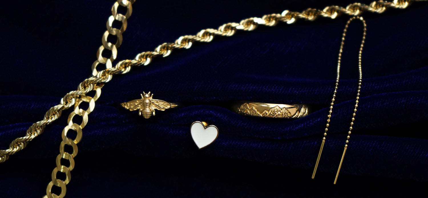 A Thick Rope Chain, Miami Chain, Bee Ring, Enamel Heart Earring, Mountains Band, and a Threader on a dark blue background