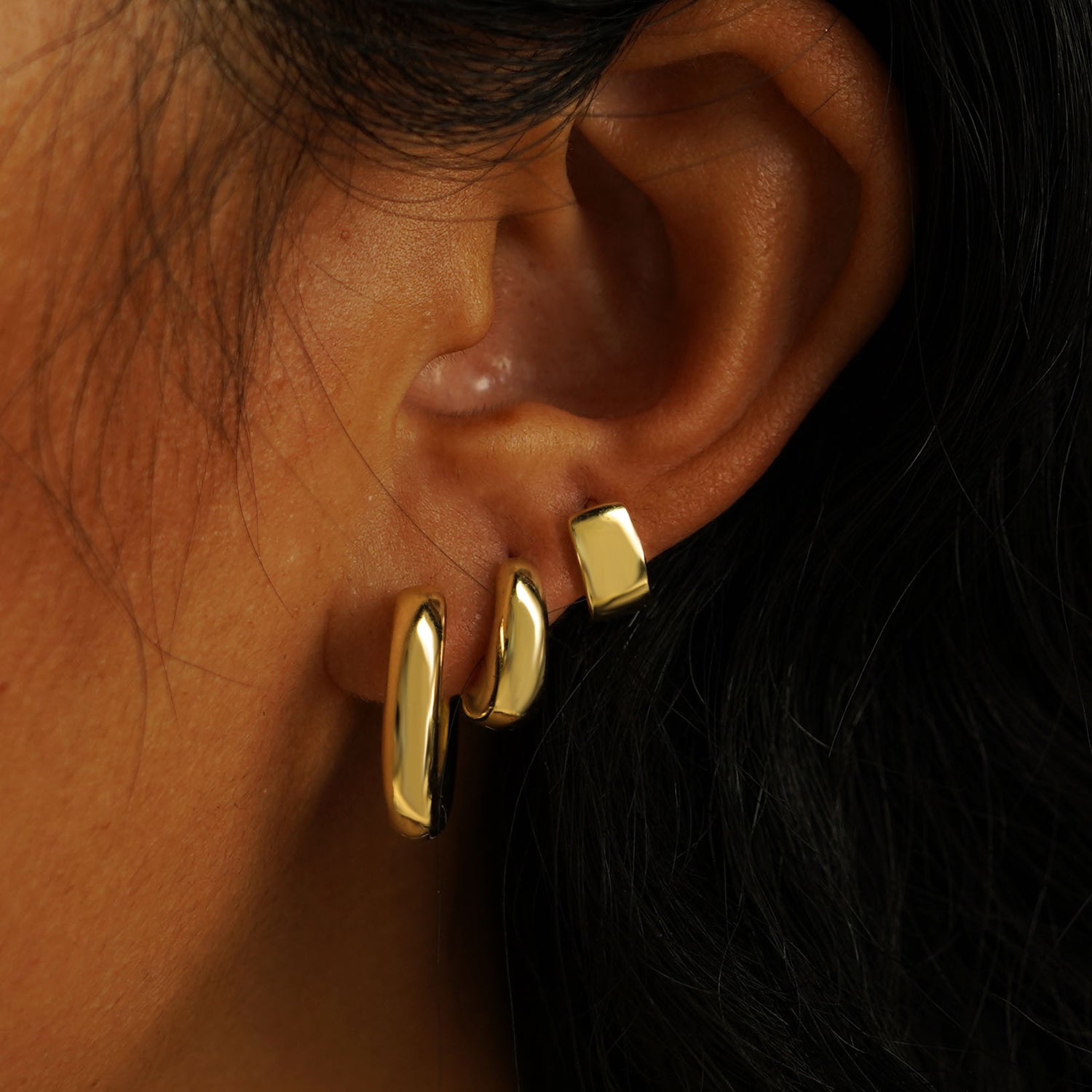 A model's ear wearing a Chunky Oval Huggie, a Small Teardrop Huggie, and an Extra Wide Huggie