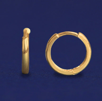 A pair of solid yellow gold Curvy Huggie Hoops on dark blue background