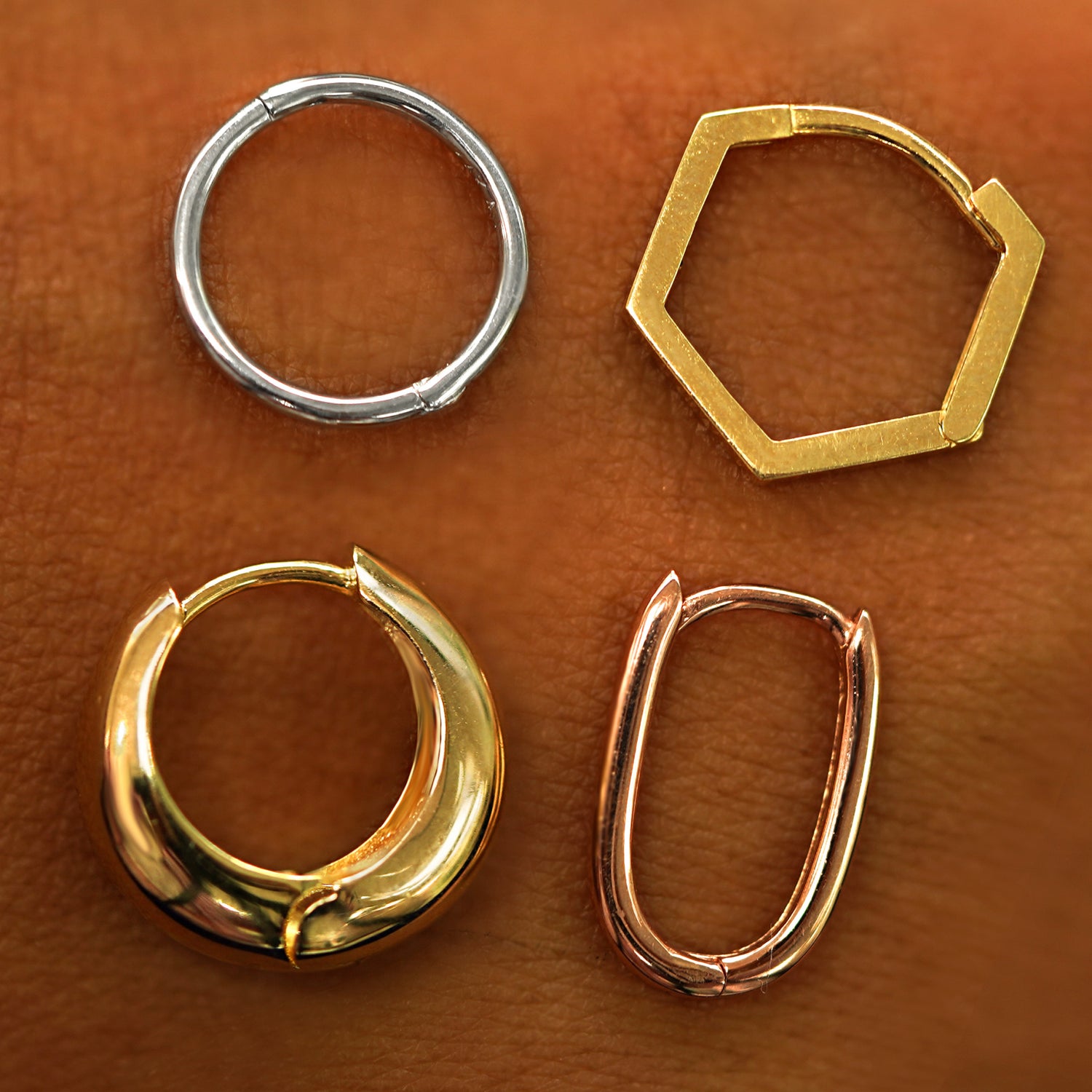 Four different geometric huggie hoops in different shades of 14k solid gold resting on the back of a model's hand