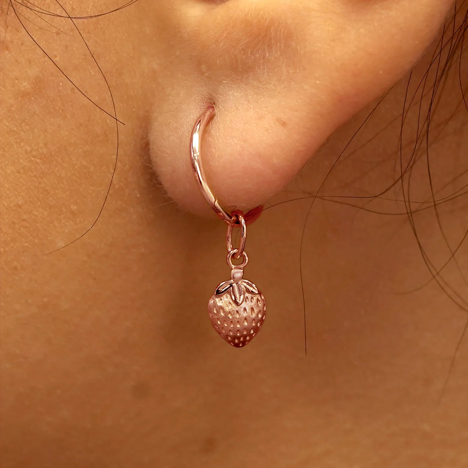 Close up view of a model's ear wearing a rose gold Strawberry Charm on a Small Seamless Huggie Hoop
