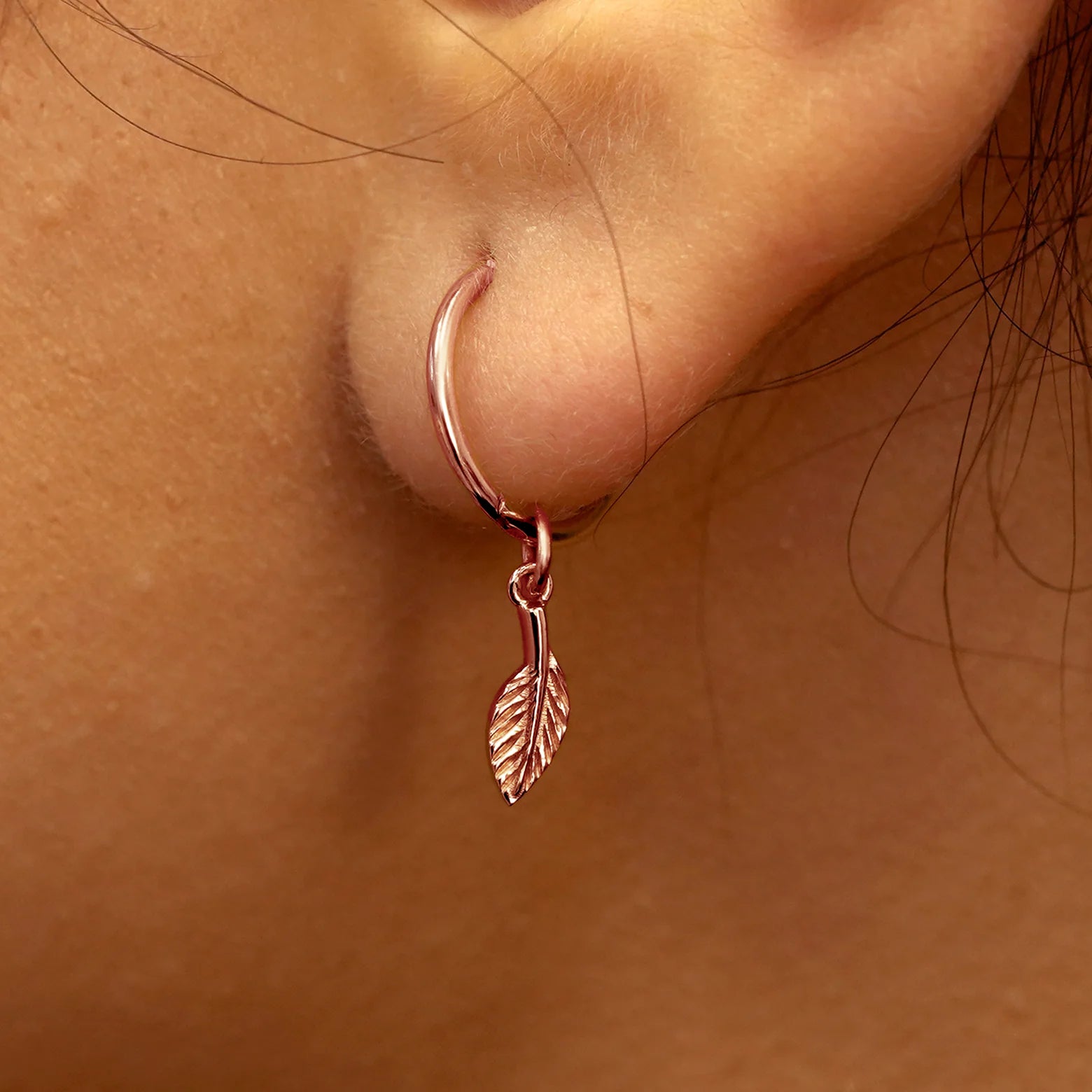 Close up view of a model's ear wearing a rose gold Leaf Charm on a Small Seamless Huggie Hoop