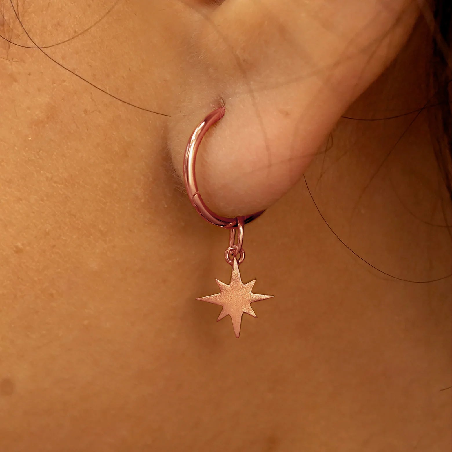 Close up view of a model's ear wearing a rose gold Starburst Charm on a Small Seamless Huggie Hoop
