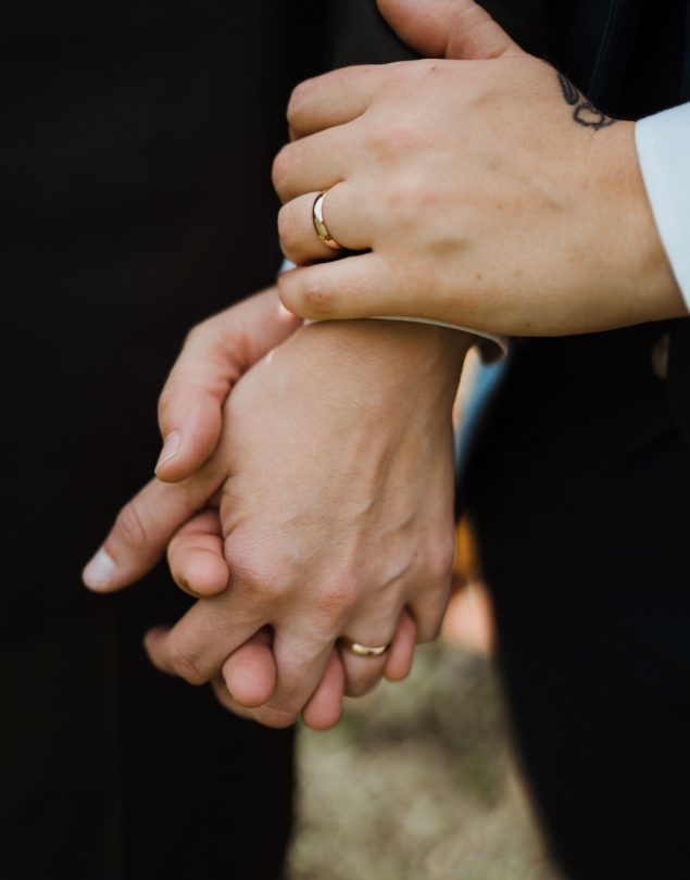 Close up view of a couple holding hands while wearing matching Automic Gold 14 karat yellow gold Curvy Mirror wedding bands