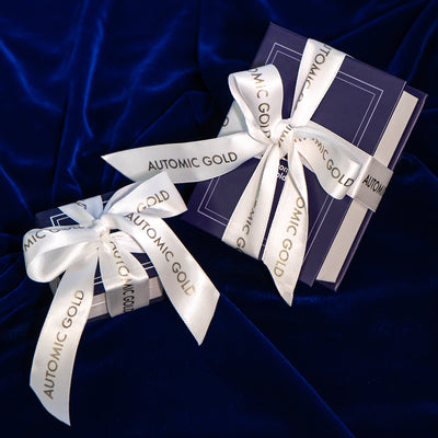 An Automic Gold Ring Box and Book Box both gift wrapped in a white ribbon that says Automic Gold in gold letters