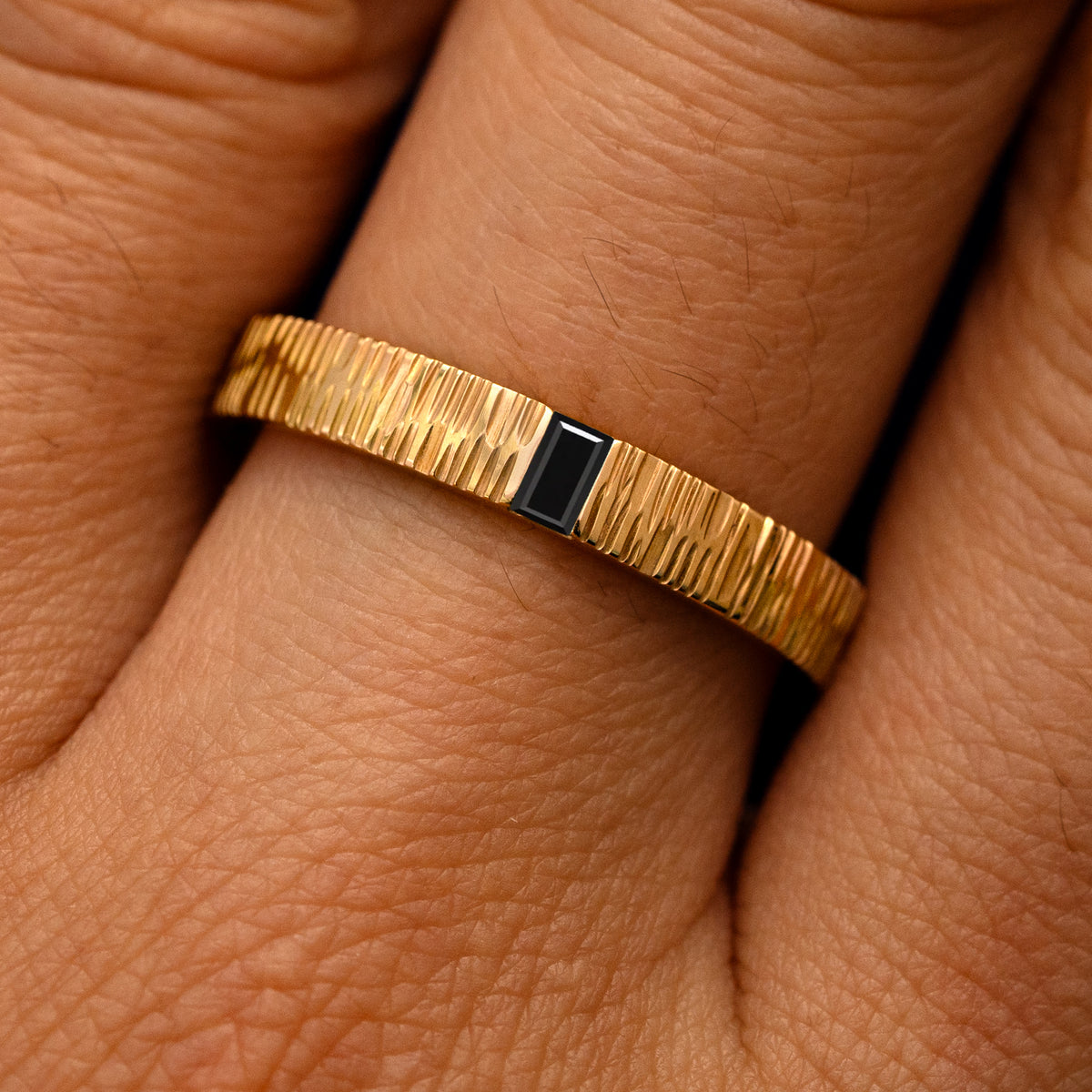 Close up view of a model's fingers wearing a 14k yellow gold Baguette Black Diamond Wood Band