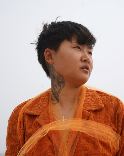 A model standing against a cloudy sky dressed in orange and wearing various Automic Gold jewelry