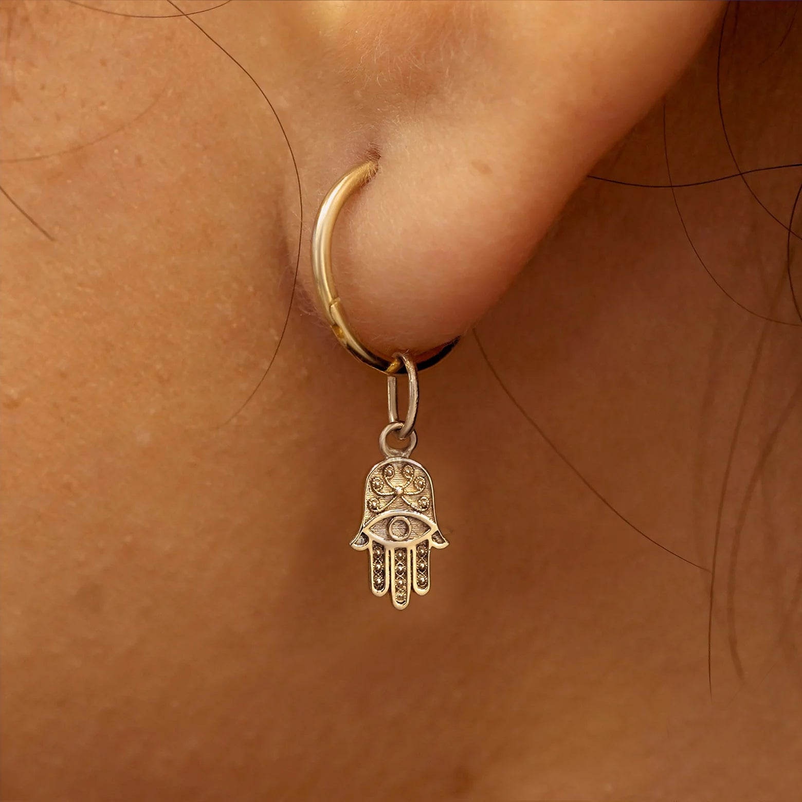 Close up view of a model's ear wearing a champagne gold Hamsa hand charm on a Small Seamless Huggie Hoop
