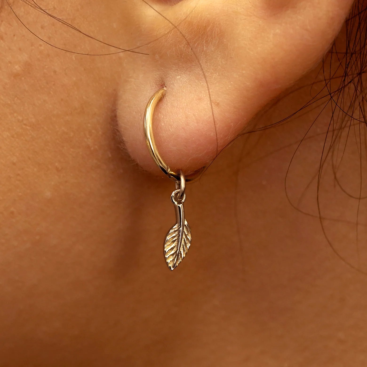 Close up view of a model's ear wearing a champagne gold Leaf Charm on a Small Seamless Huggie Hoop