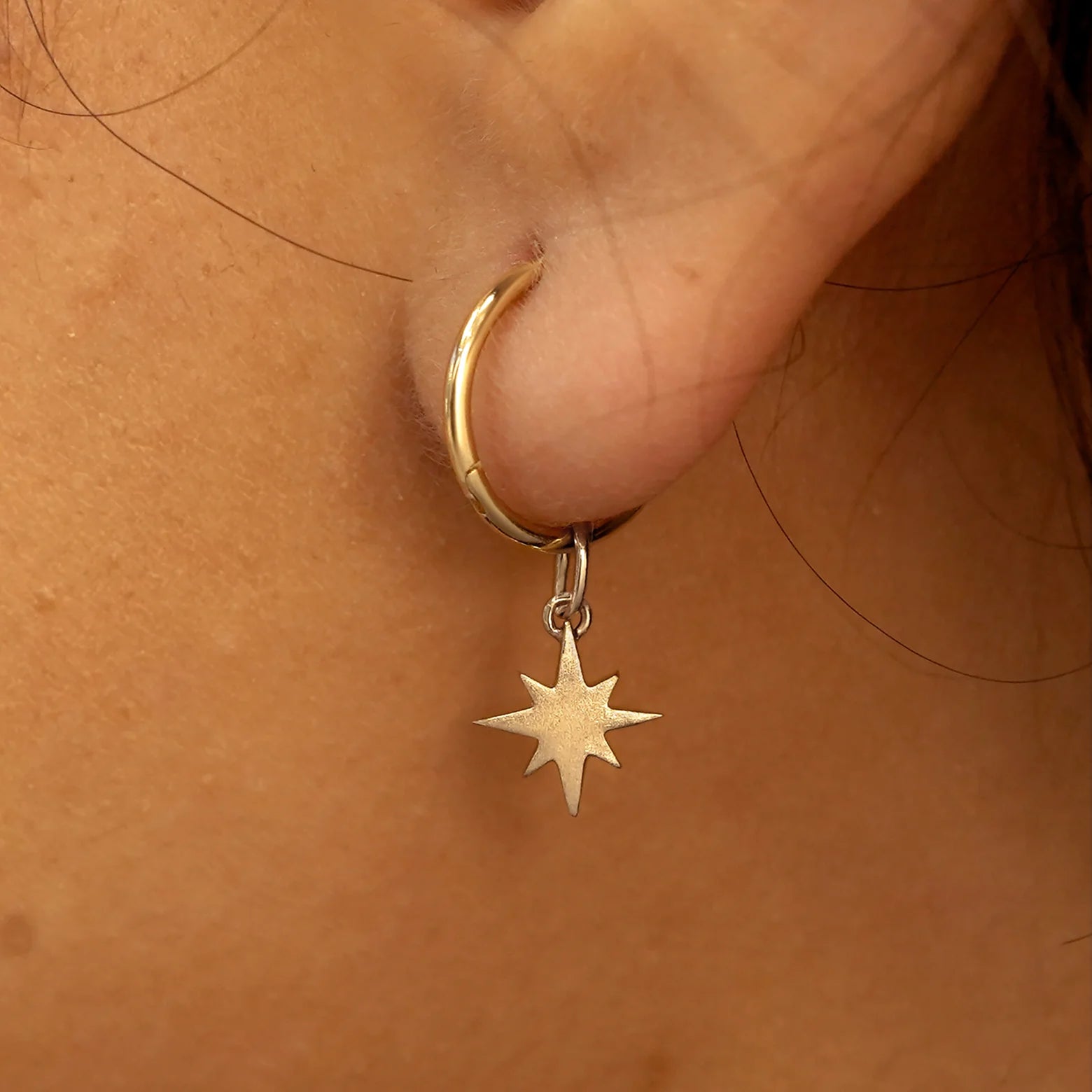 Close up view of a model's ear wearing a champagne gold Starburst Charm on a Small Seamless Huggie Hoop