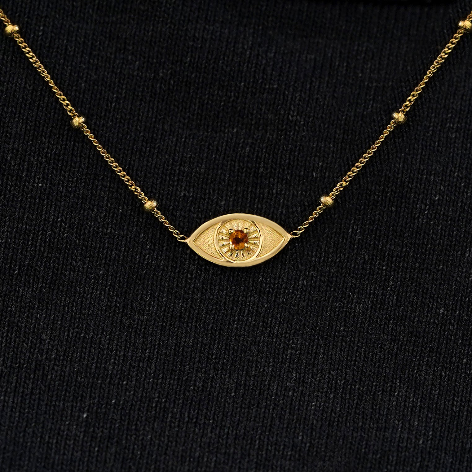 Close up view of a solid 14k yellow gold citrine evil eye necklace against a model's black shirt