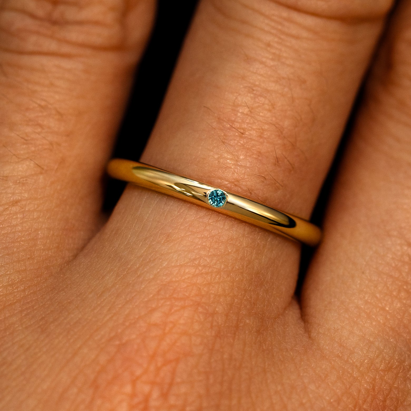 A 14k yellow gold Flush Set Round Teal Sapphire Ring on a model's finger