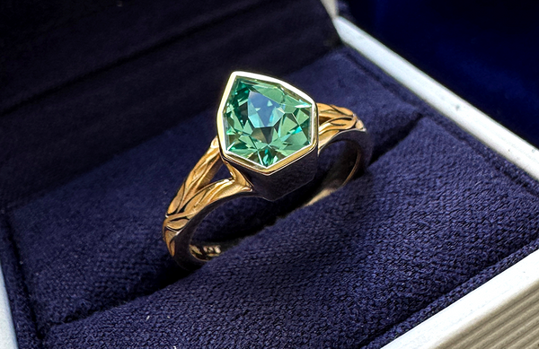 A custom ring with a shield shaped emerald in a bezel setting sits in an Automic Gold ring ox