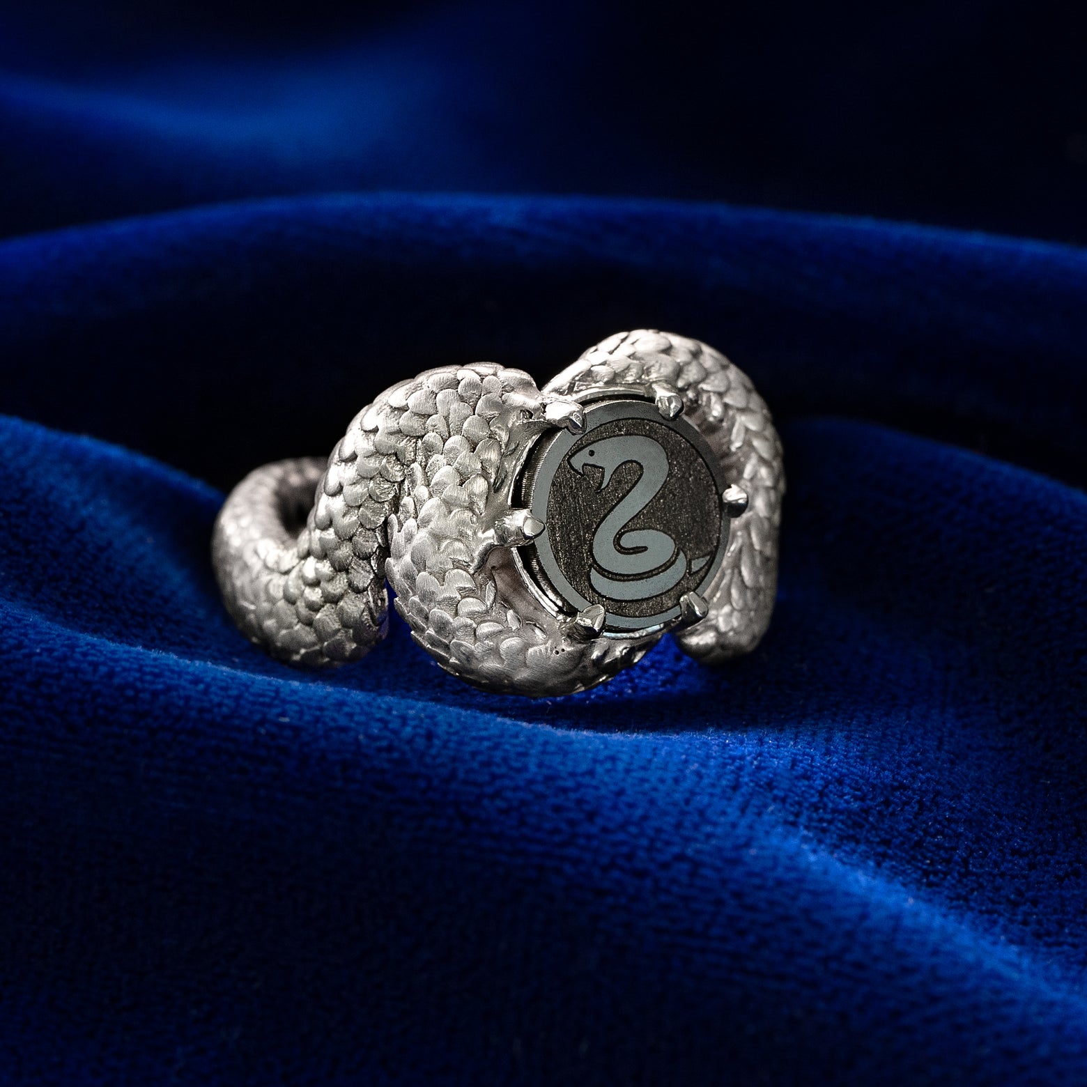 Silver ring with an engraved snake on a black diamond disc as its center on a blue fabric background