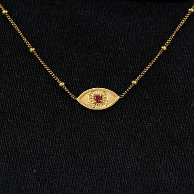 Close up view of a solid 14k yellow gold garnet evil eye necklace against a model's black shirt