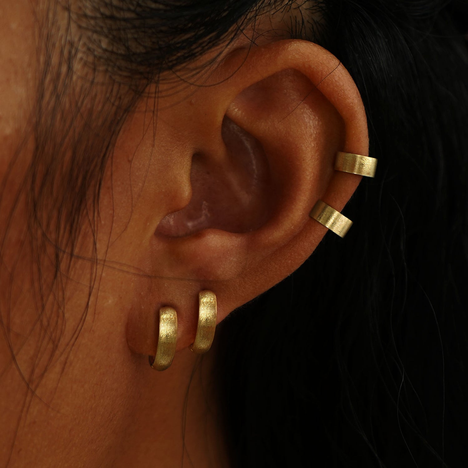 A model's ear wearing two yellow gold Stardust Thick Cuffs and two Stardust Small Wide Huggie Hoops