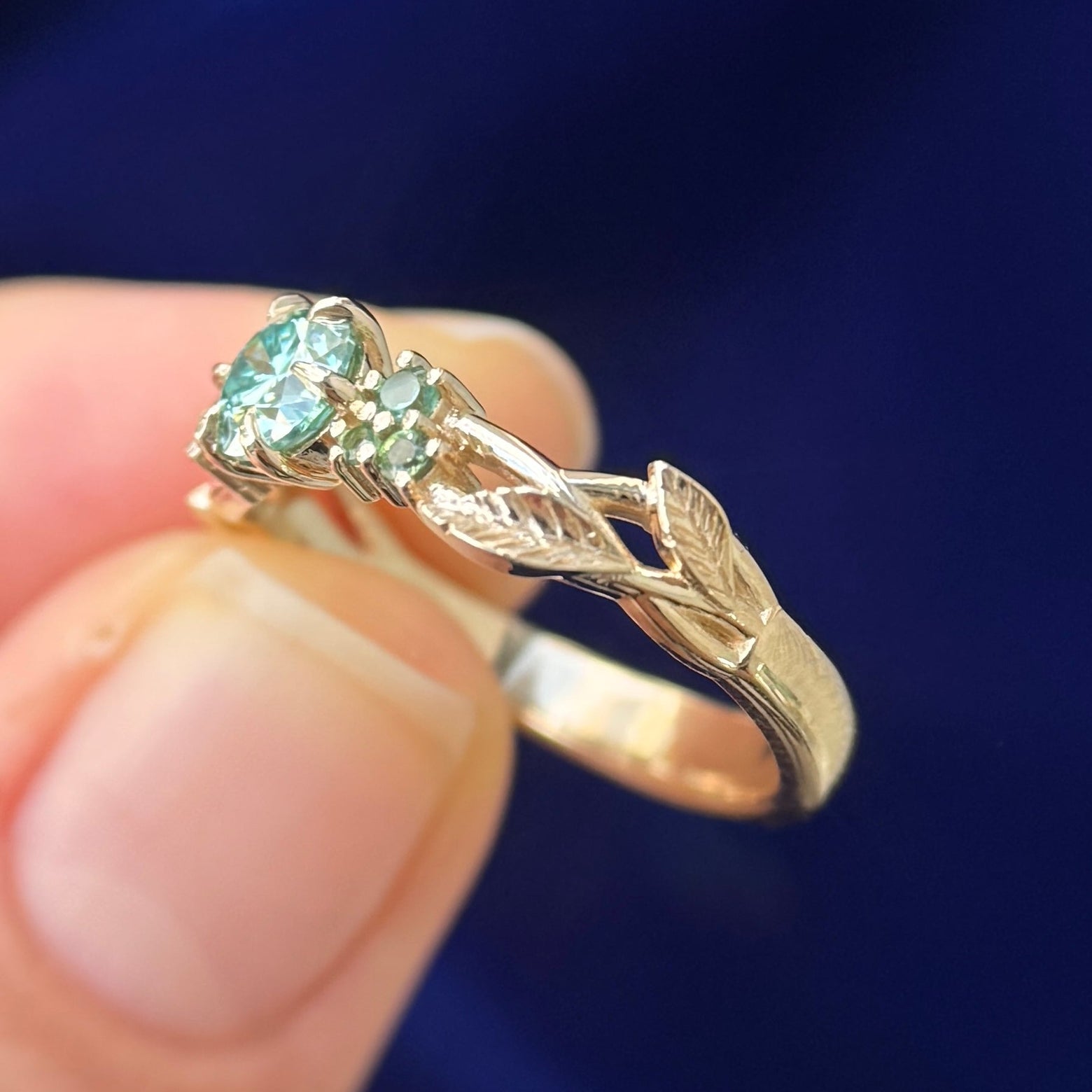 A model holding 14k Yellow Gold and 6-claw-prongs-set 5mm Round Light Green Moissanite with Green Sapphires Cluster on Leaves and Branches Custom Ring