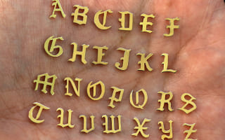 A model's palm holding solid 14k yellow gold versions of all 26 initials in the unique Automic Gold font