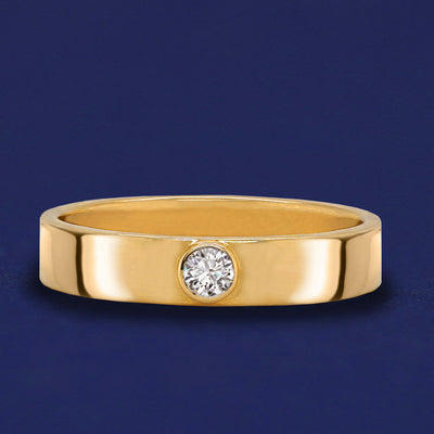 A 14 karat solid gold band ring with a bezel set Diamond on a dark blue background