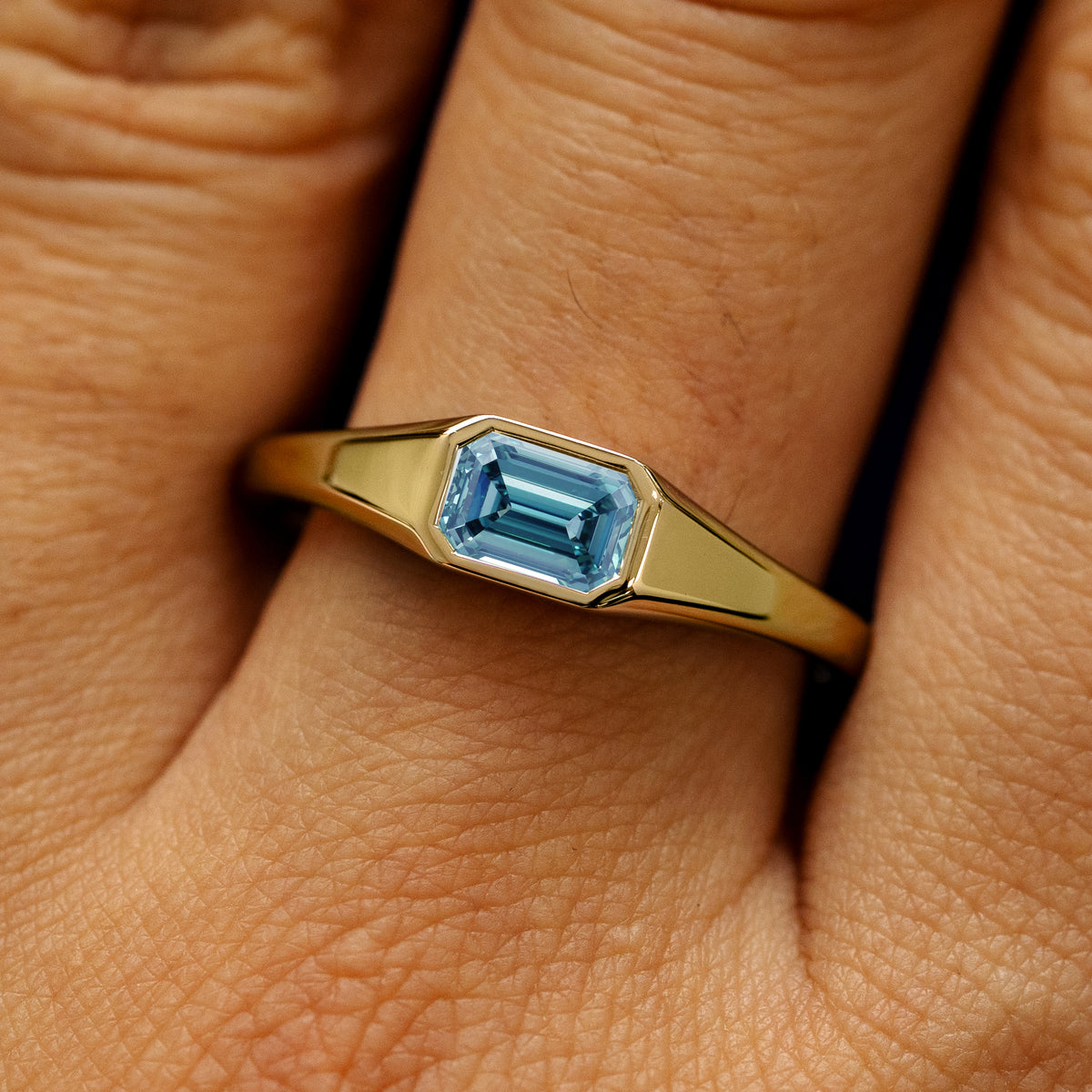 Close up view of a model's fingers wearing a 14k yellow gold emerald cut light blue lab created diamond Signet Ring