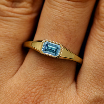 Close up view of a model's fingers wearing a 14k yellow gold emerald cut light blue lab created diamond Signet Ring