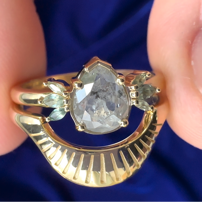 This 14k yellow gold band features and light pear salt and pepper diamond with 4 rounded prongs and 6 marquise light green sapphires with a matching band that has sunray-like engravings on the face of it. 