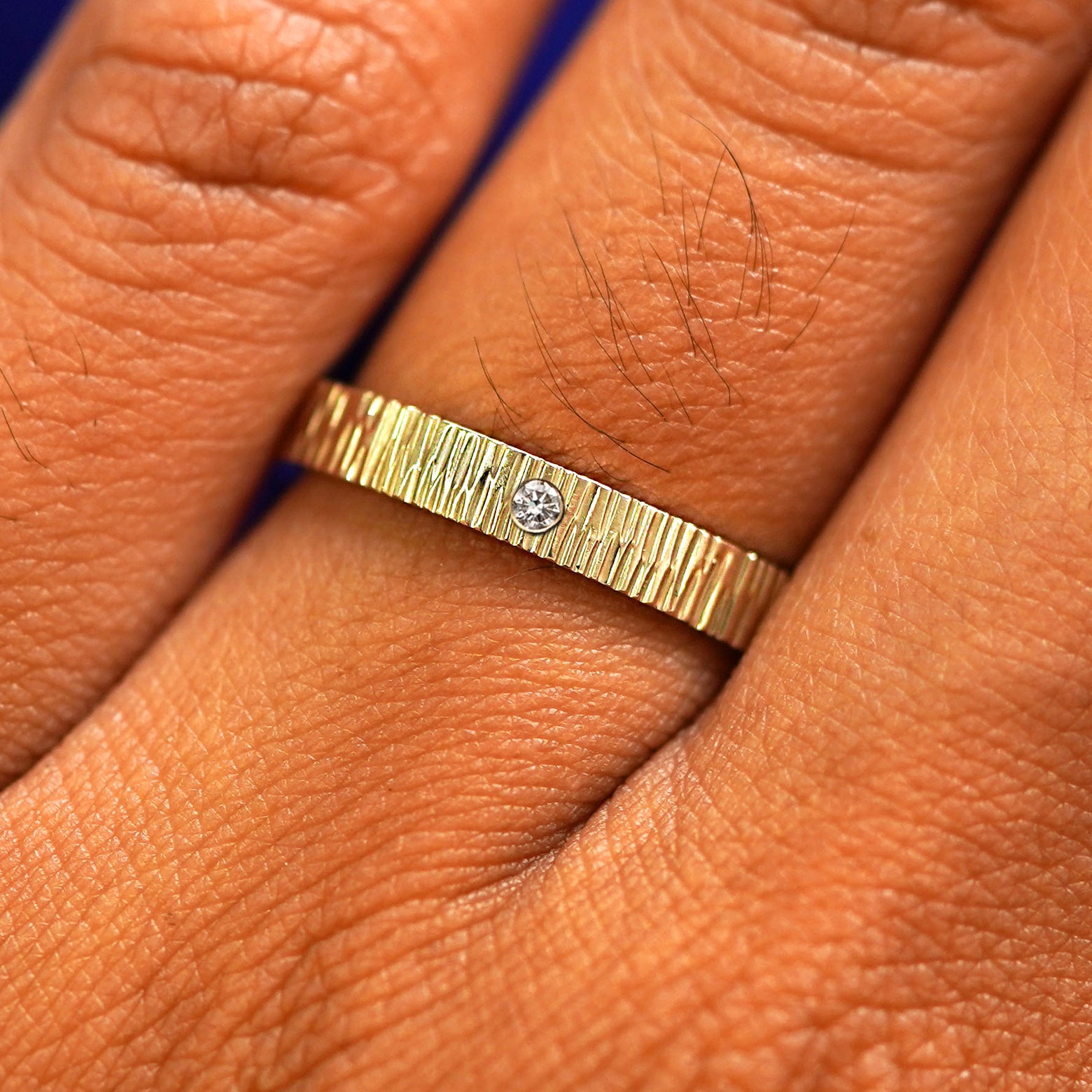 Close up view of a model's fingers wearing a 14k yellow gold Industrial Wood Band with one diamond