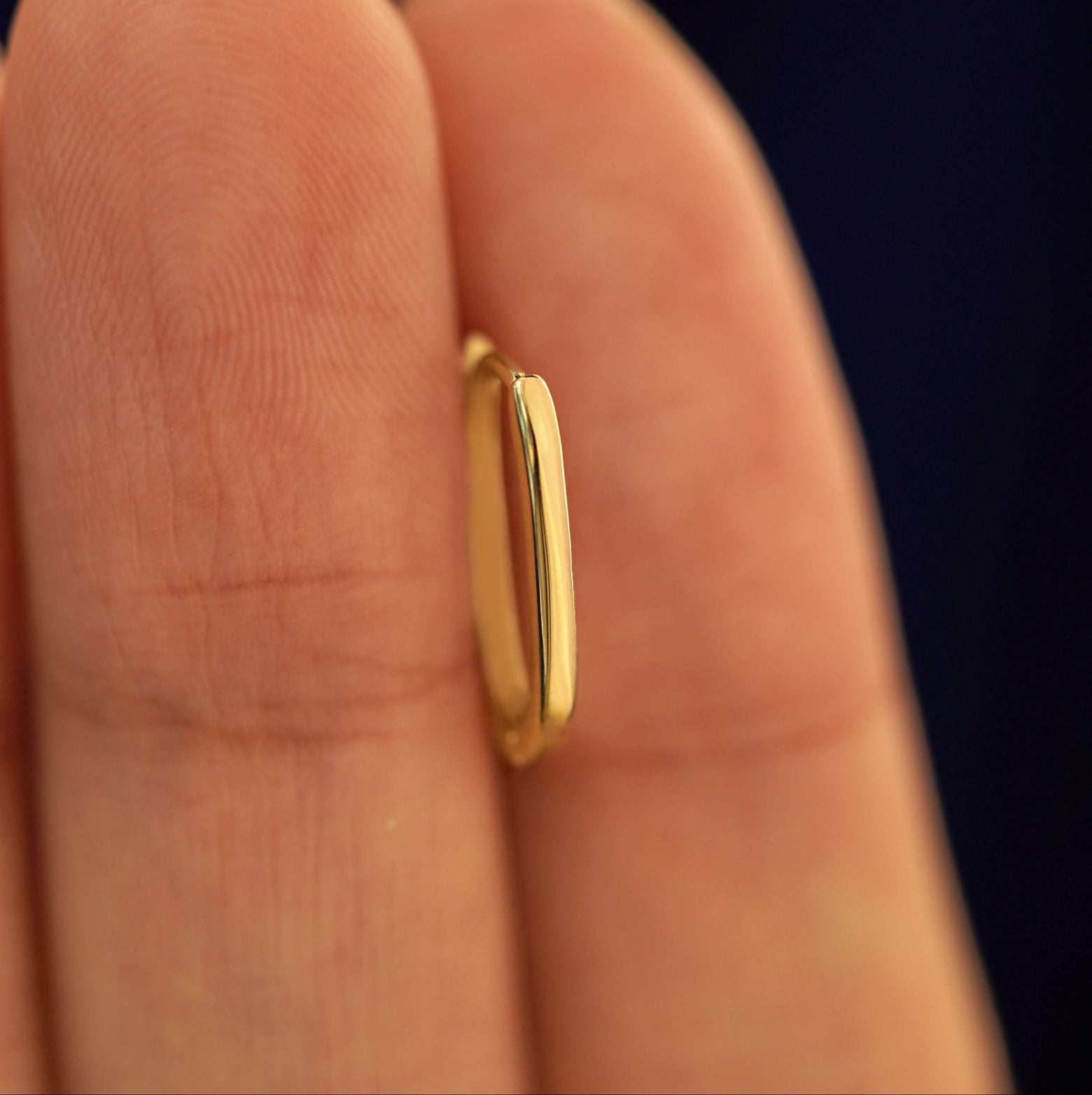 A 14k solid gold Oval Huggie Hoop between a model's fingers to show the thickness of the earring