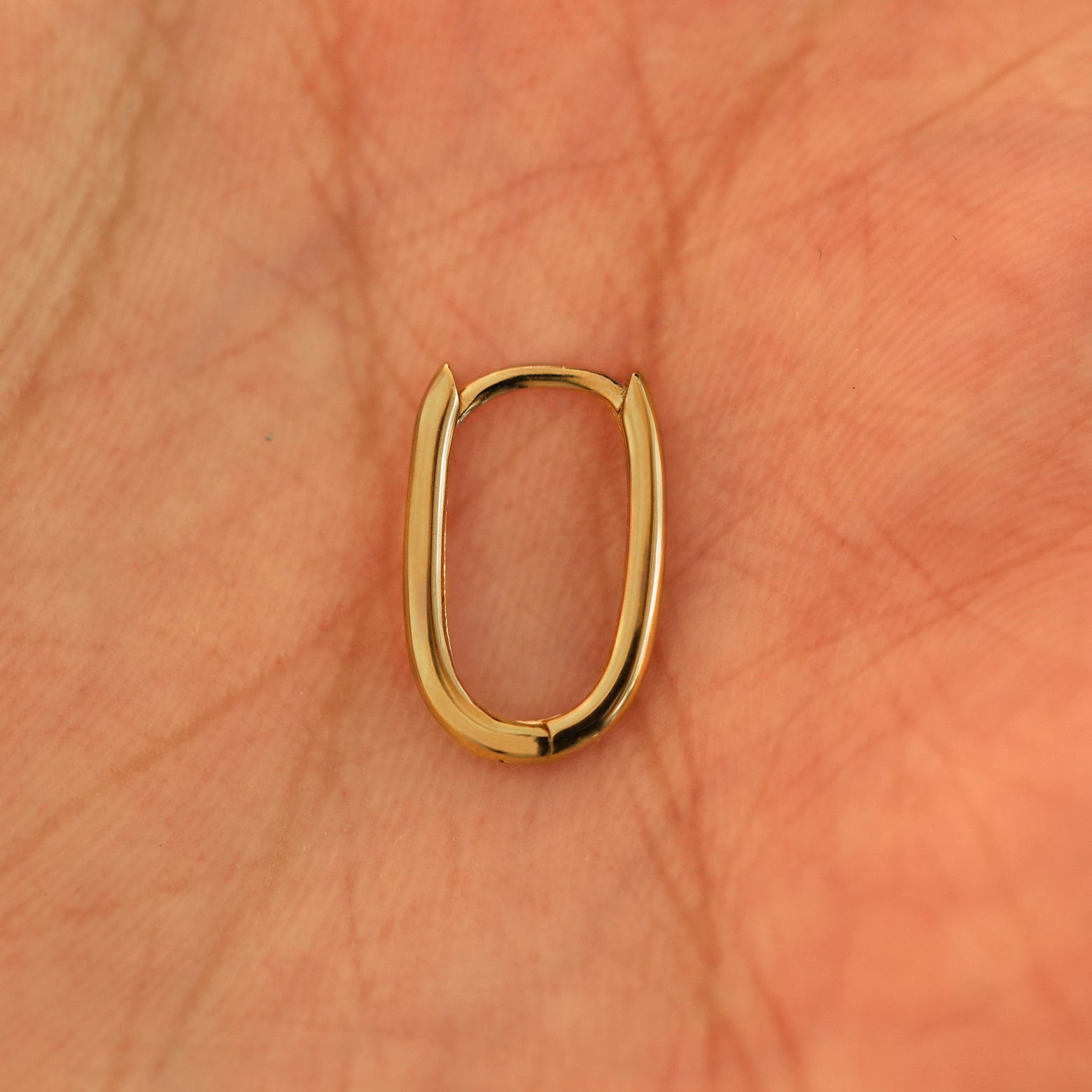 A single closed 14k yellow gold Oval Huggie Hoop resting in a model's palm