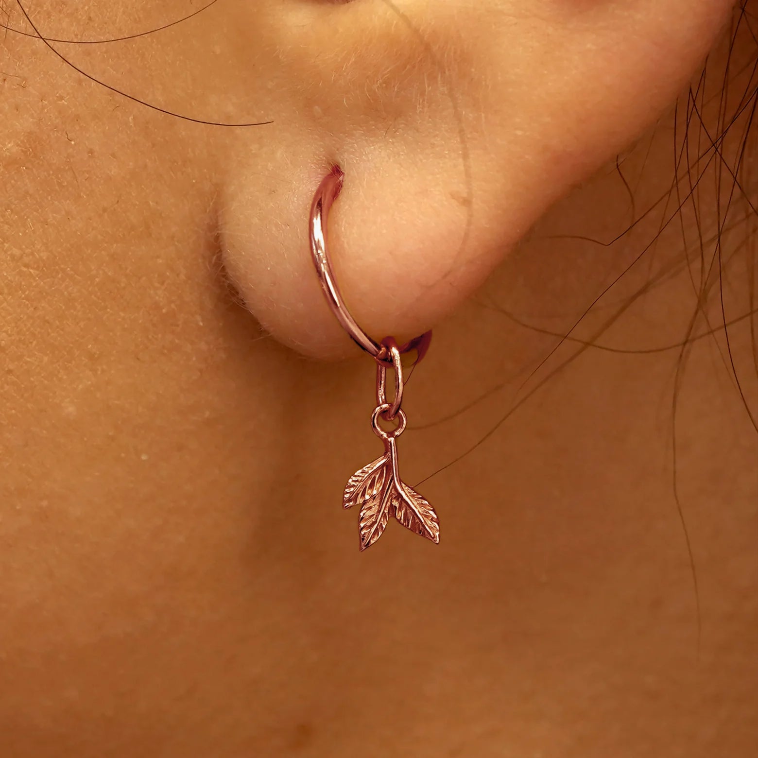 Close up view of a model's ear wearing a rose gold 3 Leaves Charm on a Small Seamless Huggie Hoop