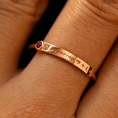A model's finger wearing a ruby champagne gold bezel set gemstone bar ring with the word engraving in cursive font