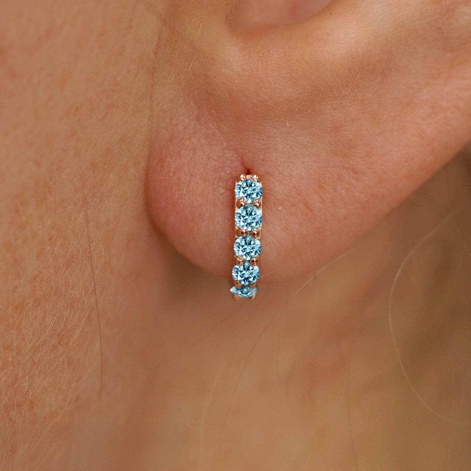 Close up view of a model's ear wearing a rose gold Mini Light Blue Lab Diamond Huggie Hoop