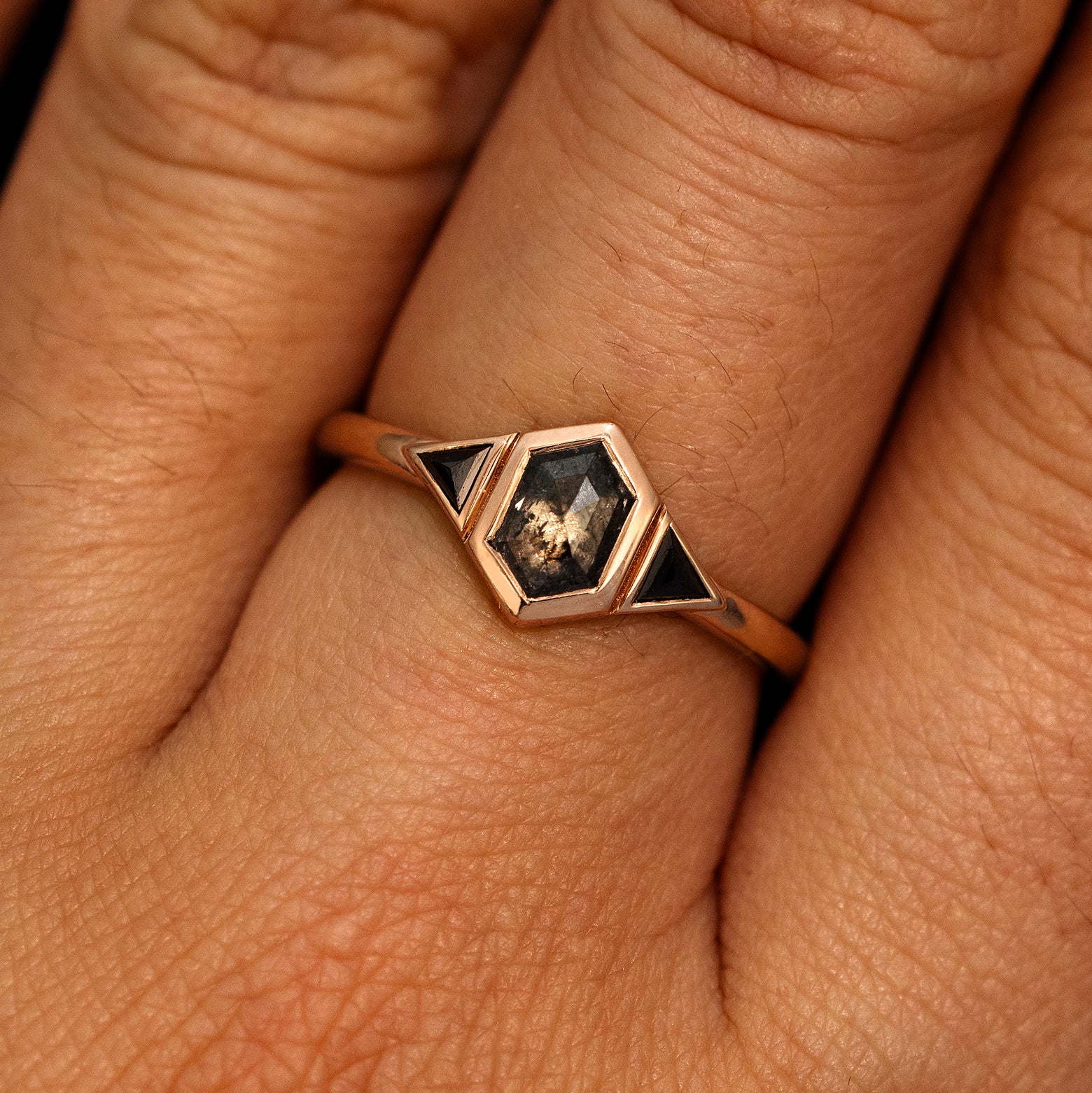 A model's hand wearing a 14k rose gold Hexagon Salt and Pepper and Triangle Black Diamond Three Stone Ring