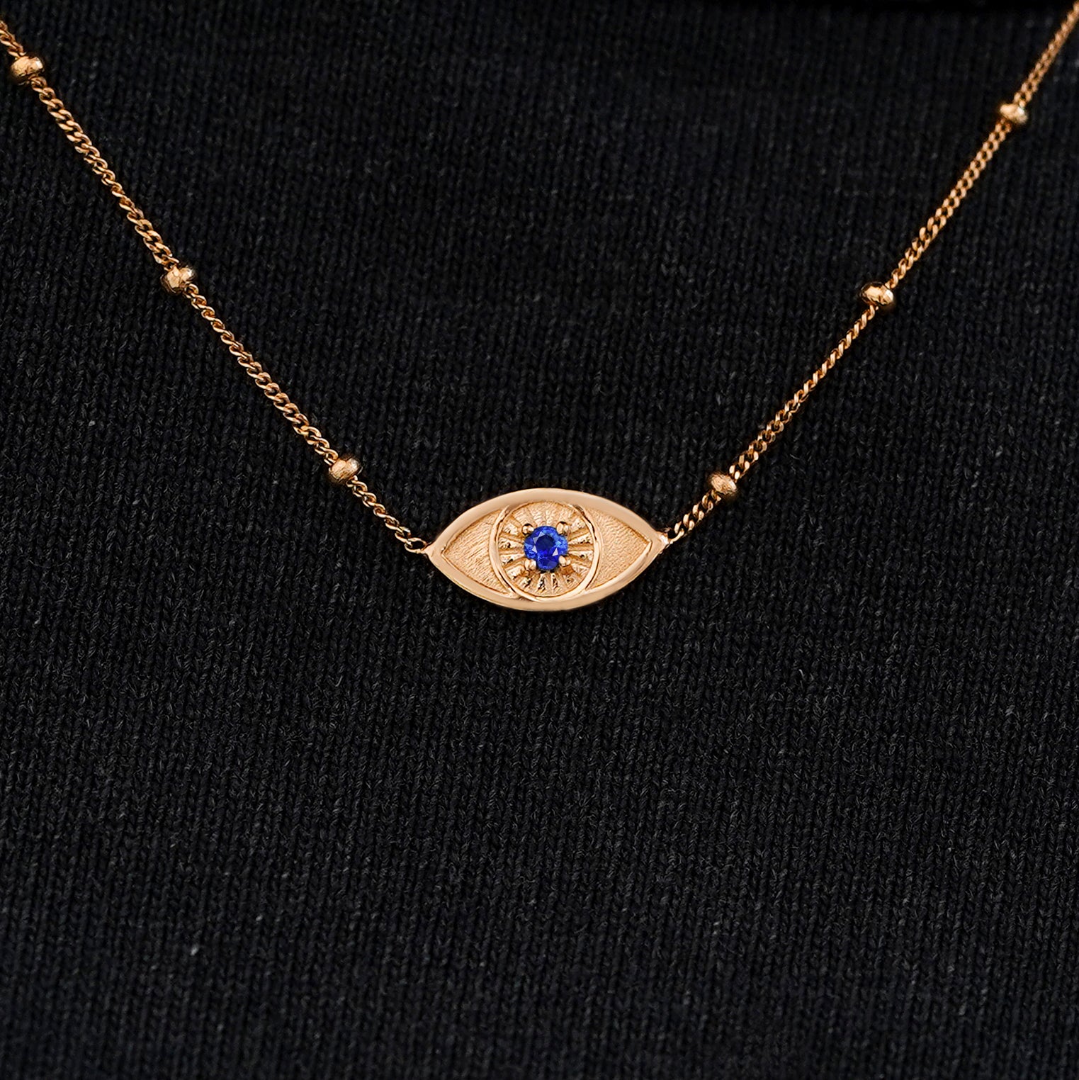 Close up view of a solid 14k rose gold sapphire evil eye necklace against a model's black shirt