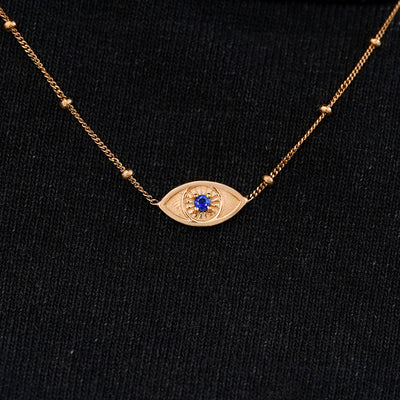Close up view of a solid 14k rose gold sapphire evil eye necklace against a model's black shirt