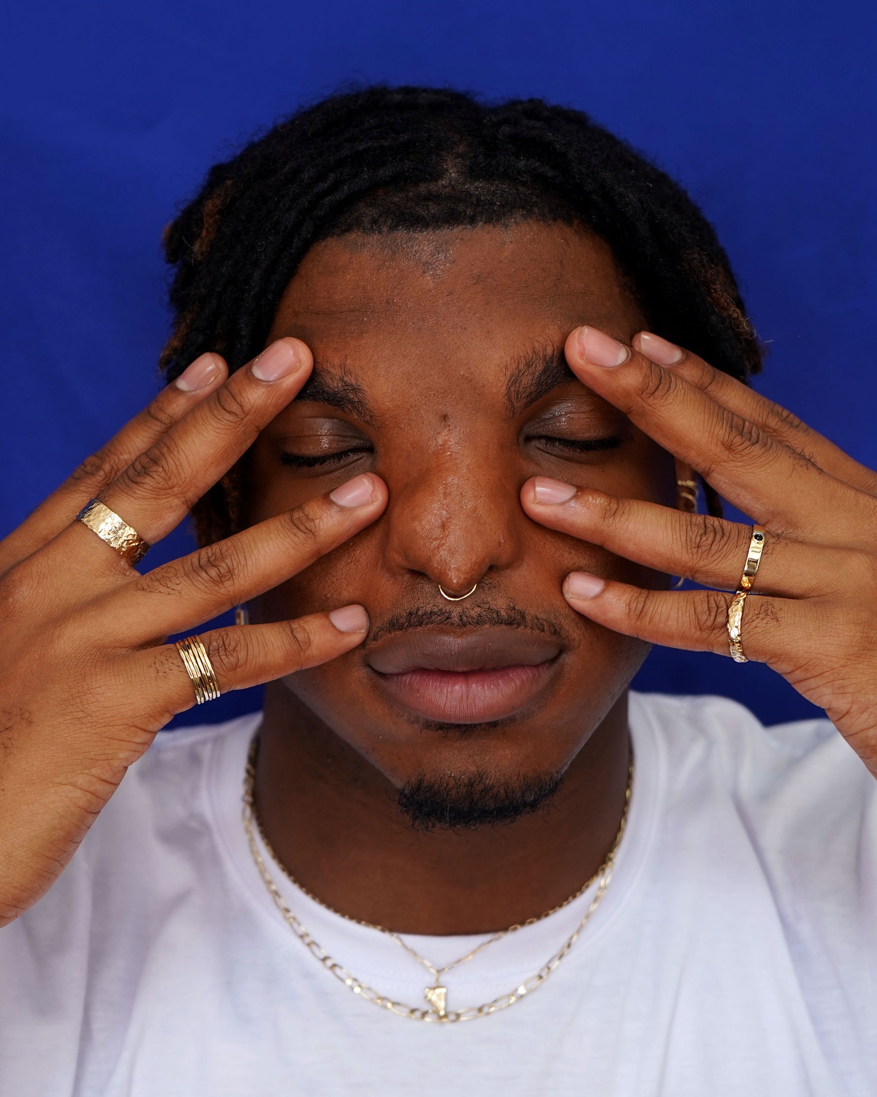 A model with both hands gently touching around their eyes wearing yellow gold rings, earrings, and a septum ring