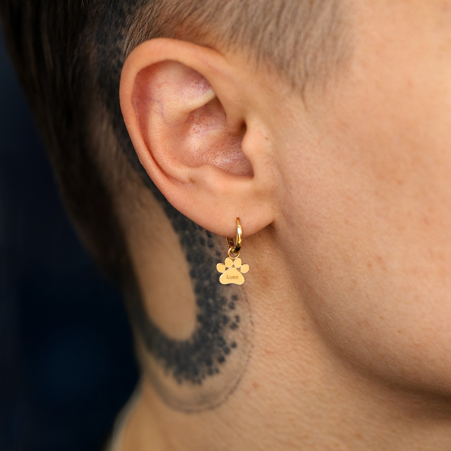 Close up view of a models ear wearing a solid gold paw charm with the name Luna engraved on it on a curvy huggie hoop earring