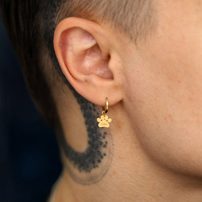 Close up view of a models ear wearing a solid gold paw charm with the name Luna engraved on it on a curvy huggie hoop earring