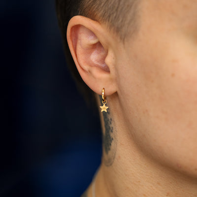 Close up view of a models ear wearing a solid gold star charm with theinitial M engraved on it on a curvy huggie hoop earring