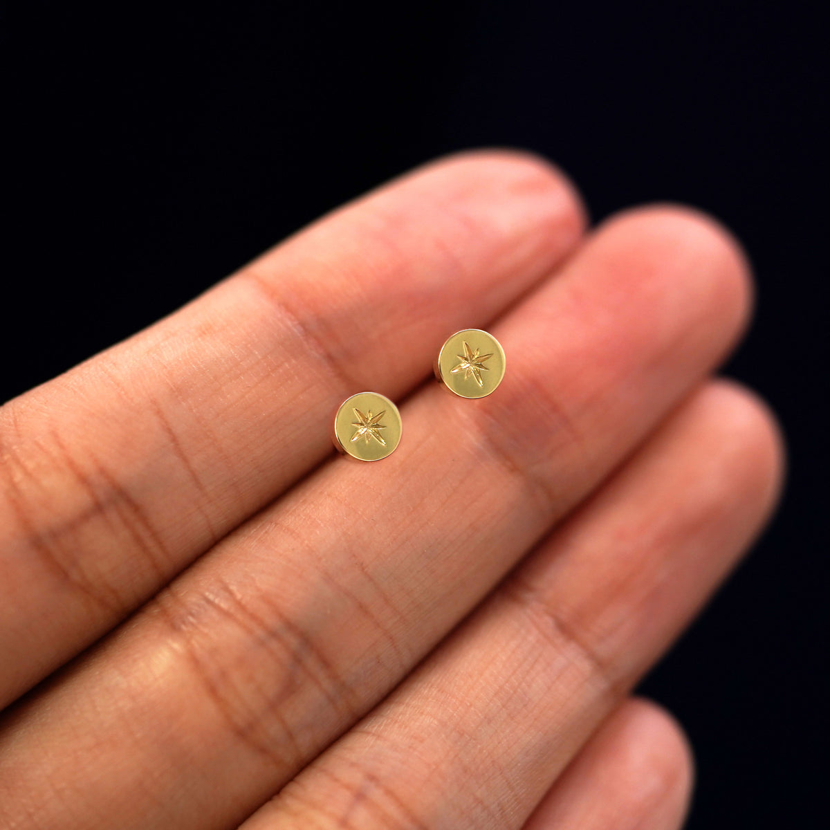 A pair of engrtaved star disk stud earrings held between a models fingers