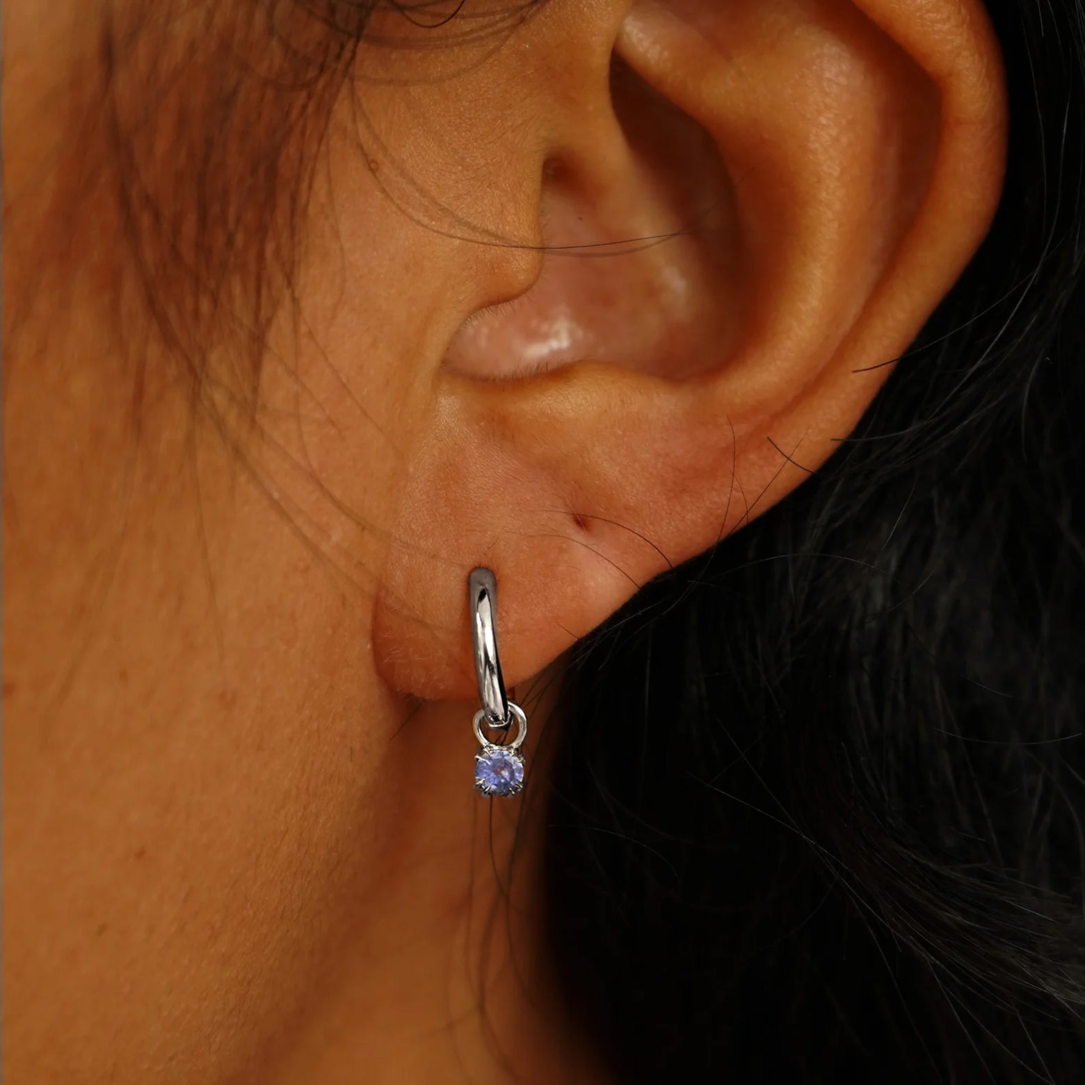 Close up view of a model's ear wearing a white gold Tanzanite Charm on a Curvy Huggie Hoop