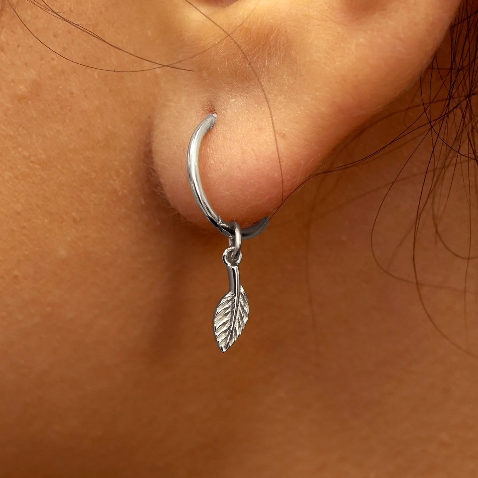 Close up view of a model's ear wearing a white gold Leaf Charm on a Small Seamless Huggie Hoop
