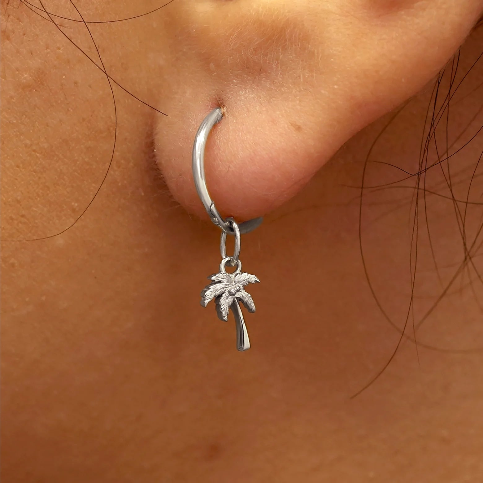 Close up view of a model's ear wearing a white gold Palm Tree Charm on a Small Seamless Huggie Hoop