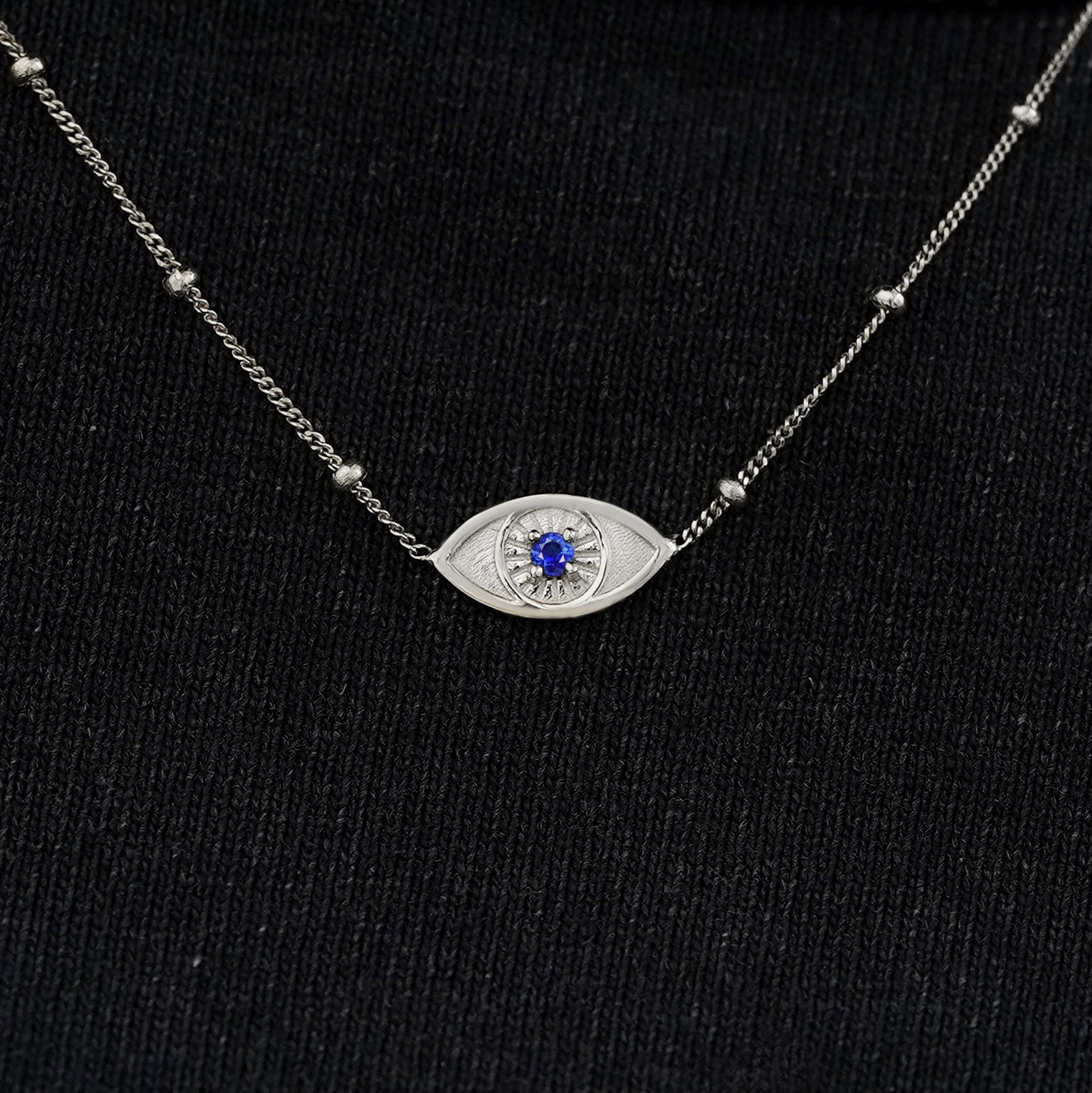 Close up view of a solid 14k white gold sapphire evil eye necklace against a model's black shirt