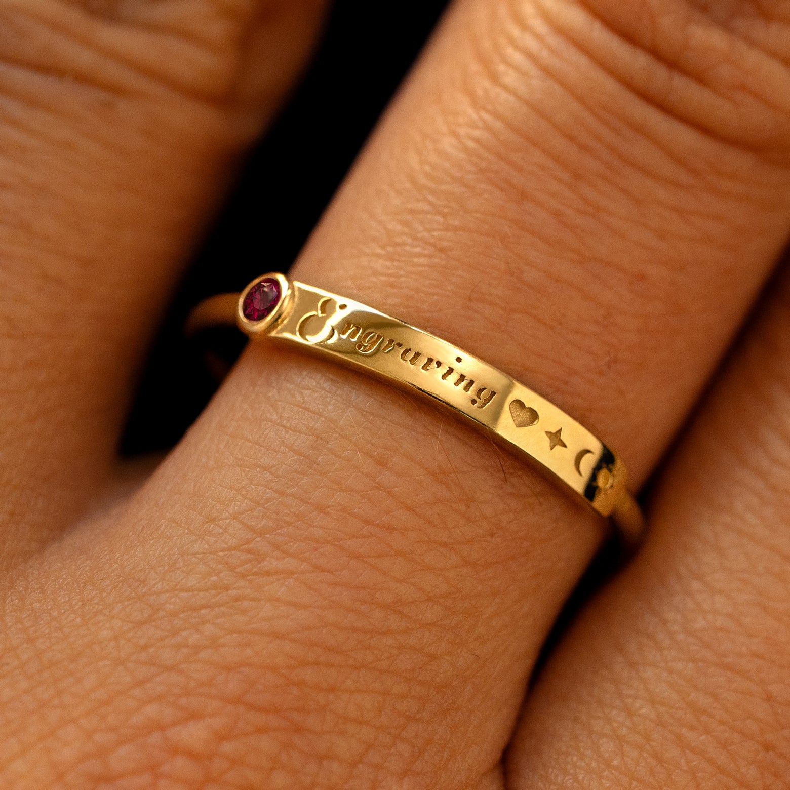 A model's finger wearing a ruby yellow gold bezel set gemstone bar ring with the word engraving in cursive font