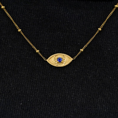 Close up view of a solid 14k yellow gold sapphire evil eye necklace against a model's black shirt