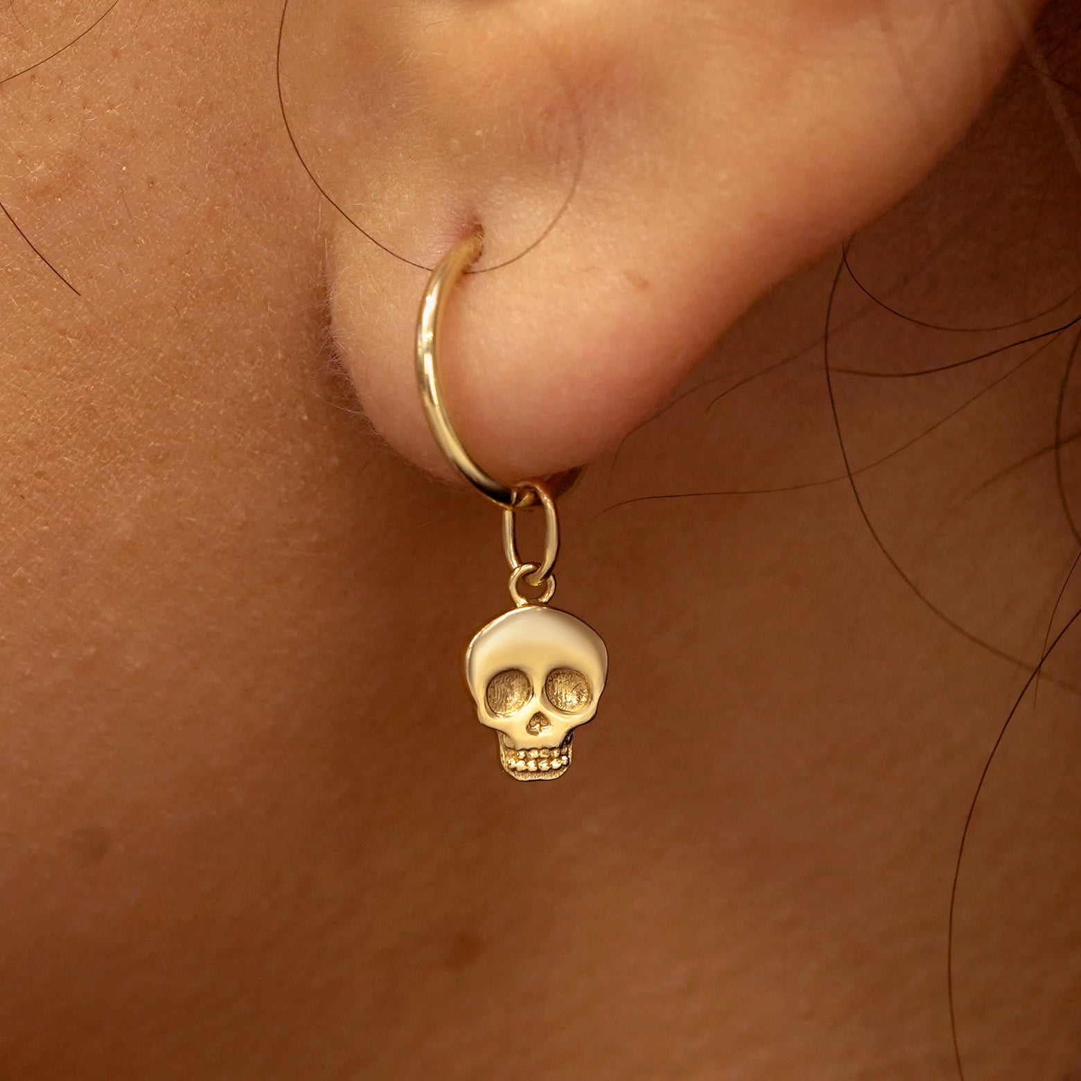 Close up view of a model's ear wearing a yellow gold Skull Charm on a Small Seamless Huggie Hoop